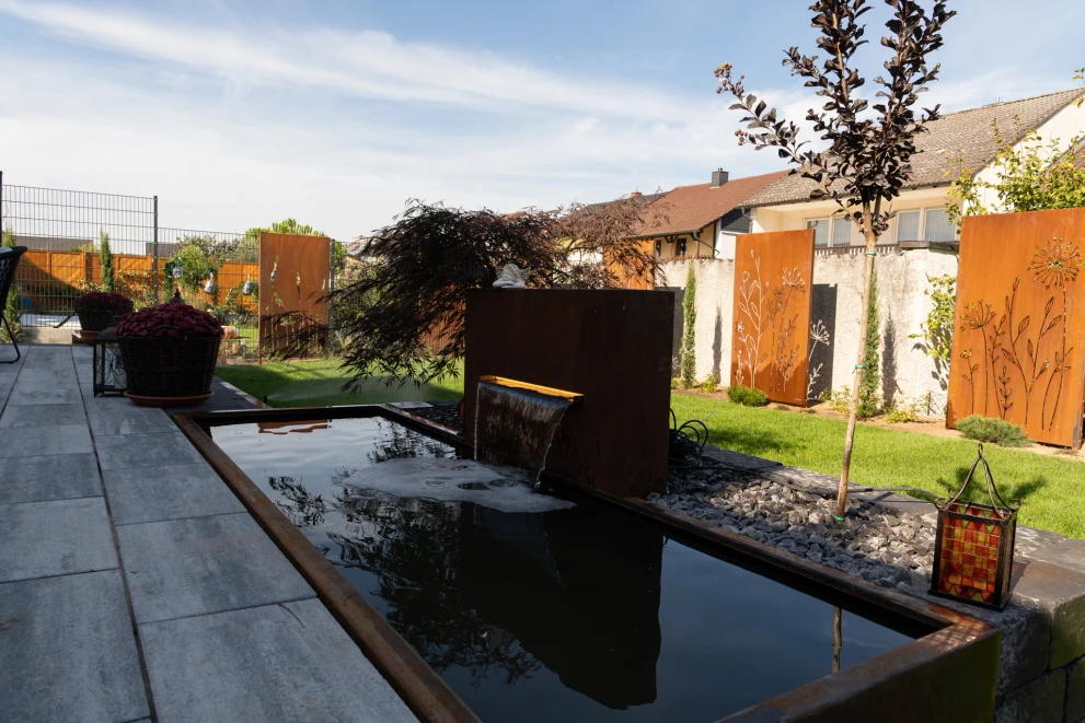 Gartenbrunnen mit breitem Wasserfall neben der Terrasse und bepflanztem Steinbeet